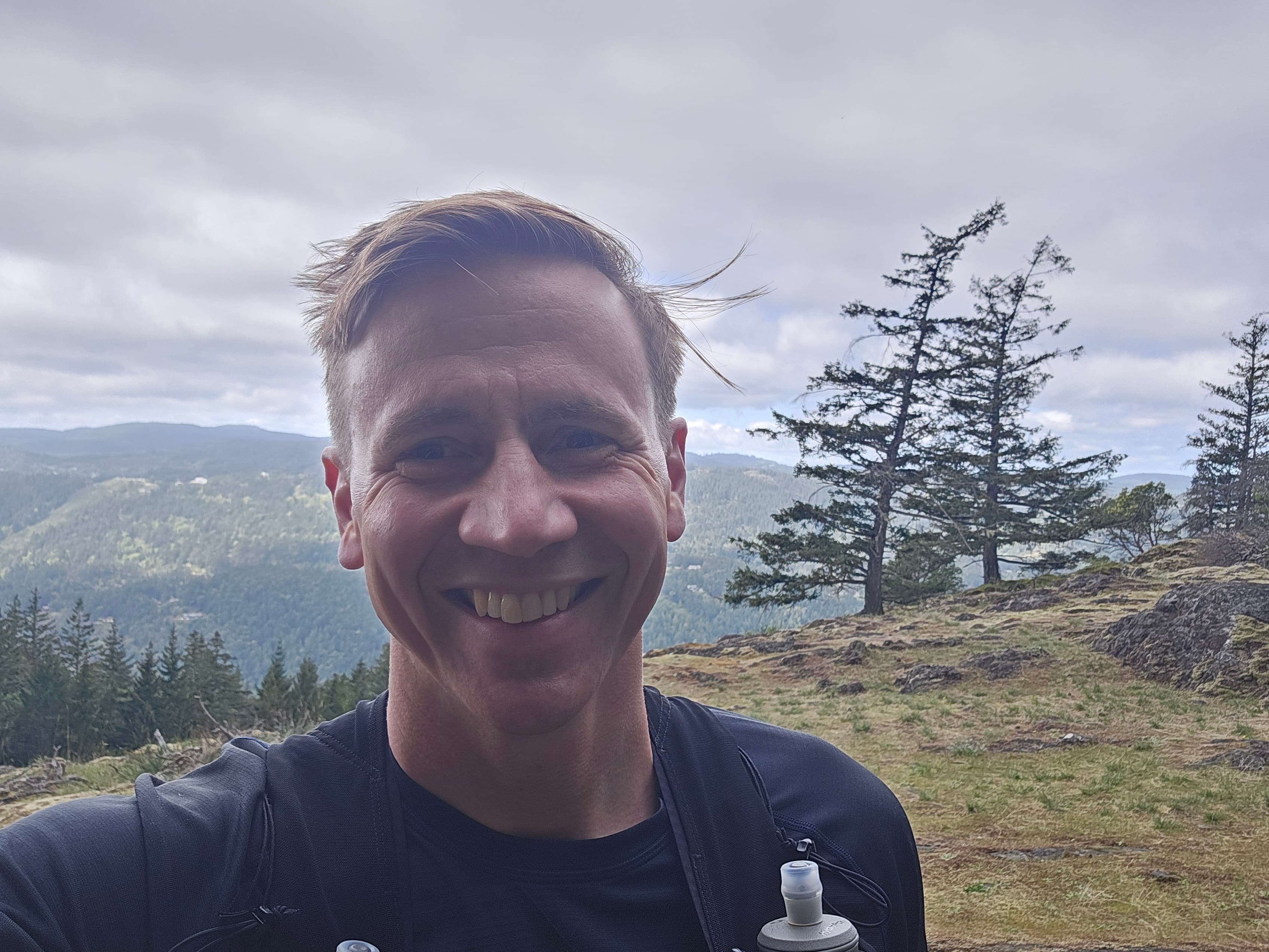 Portrait of JME Maxwell, close-up smile, mountains and forest in the background, wind in his hair.