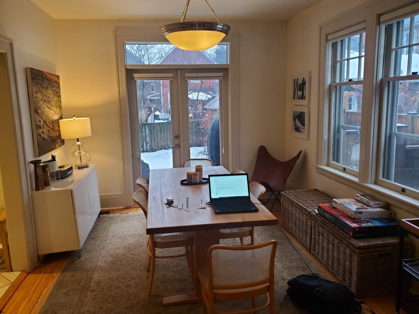 A laptop open on a wooden dining table, papers mid-task, warm pendant light, snow through the French doors.